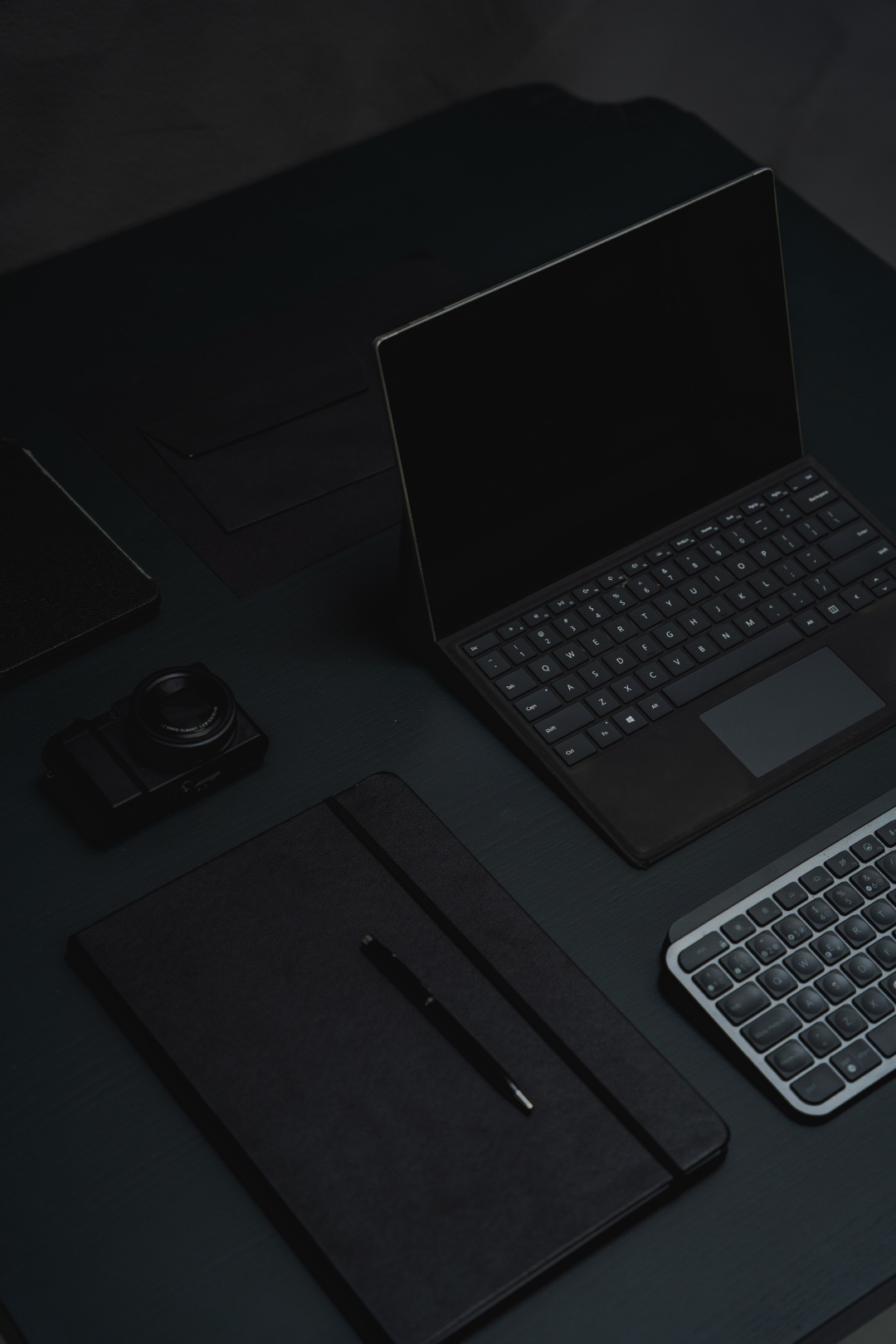 A dark, minimal desk setup with a laptop, camera, and notebook
